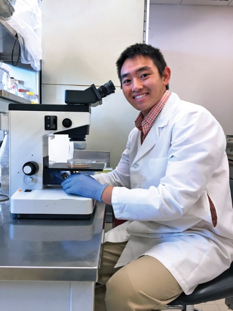 A young Asian man in a lab coat smiling while sitting at a microscope