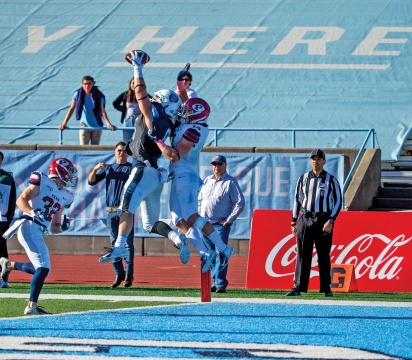 Columbia Lions football player in the air holding a football, with an opposing player behind him, also in the air, reaching for the ball.